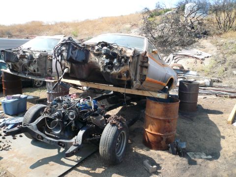 1975 Ford Ranchero - A disassembled 1975 Ford Ranchero, with its body and chassis separated and supported by barrels, sits in a junkyard.