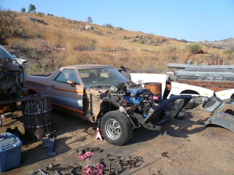1975 Ford Ranchero - A disassembled Ford Ranchero sits in a junkyard with its engine exposed and front end removed.
