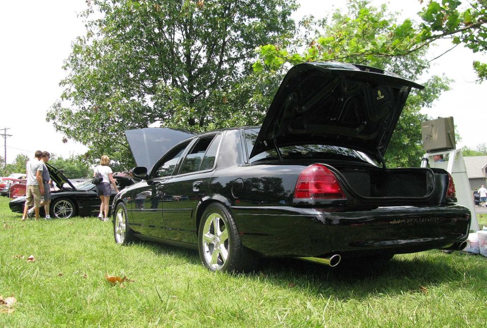 Black sedan, hood and trunk open, parked on grass with people around. Another car is visible in the background