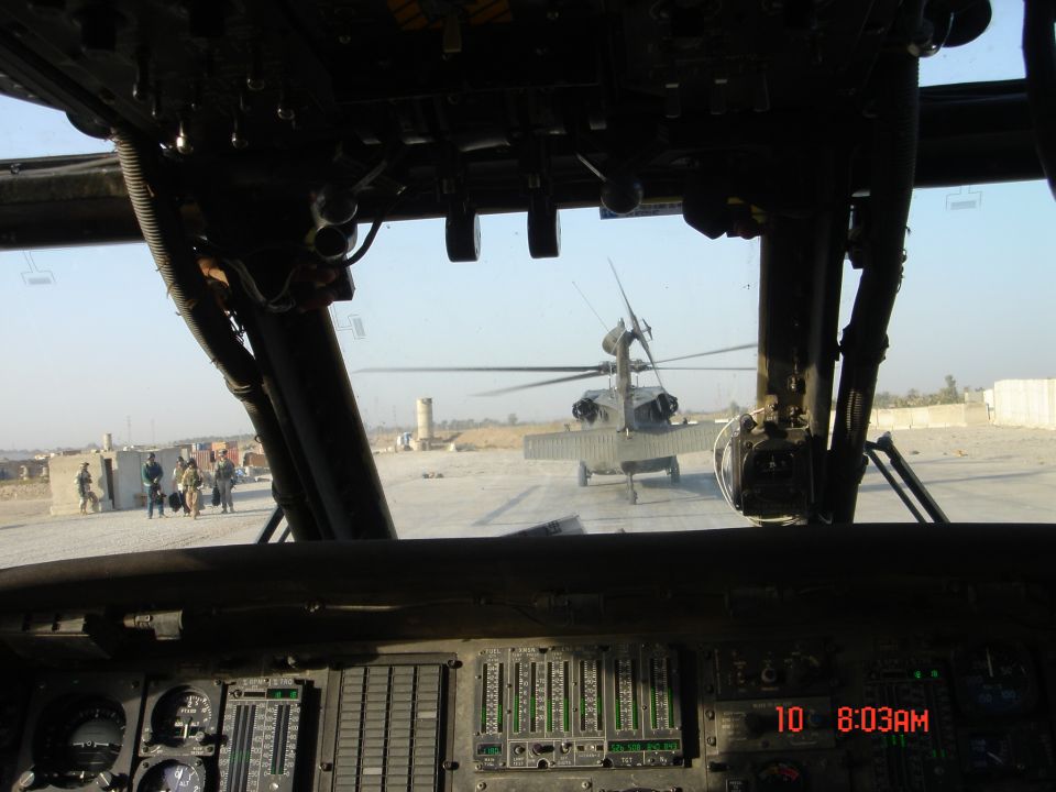 Helicopter cockpit view of a Black Hawk helicopter on a dusty airfield with instrument panel in foreground.