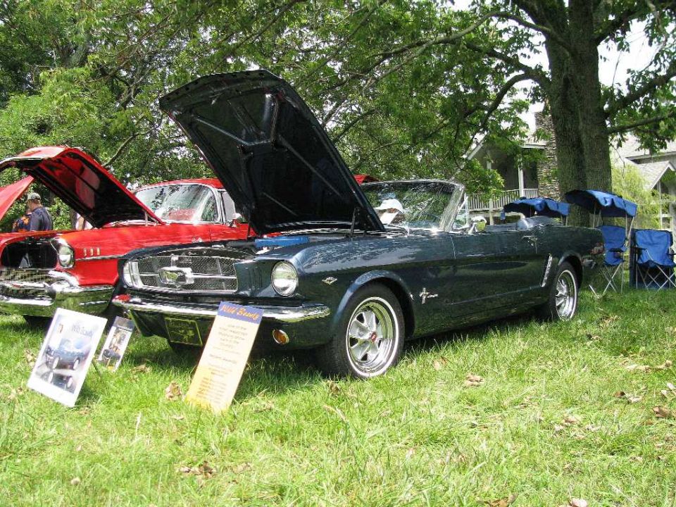 Dark green 1965 Ford Mustang convertible with hood open, parked next to a red classic car