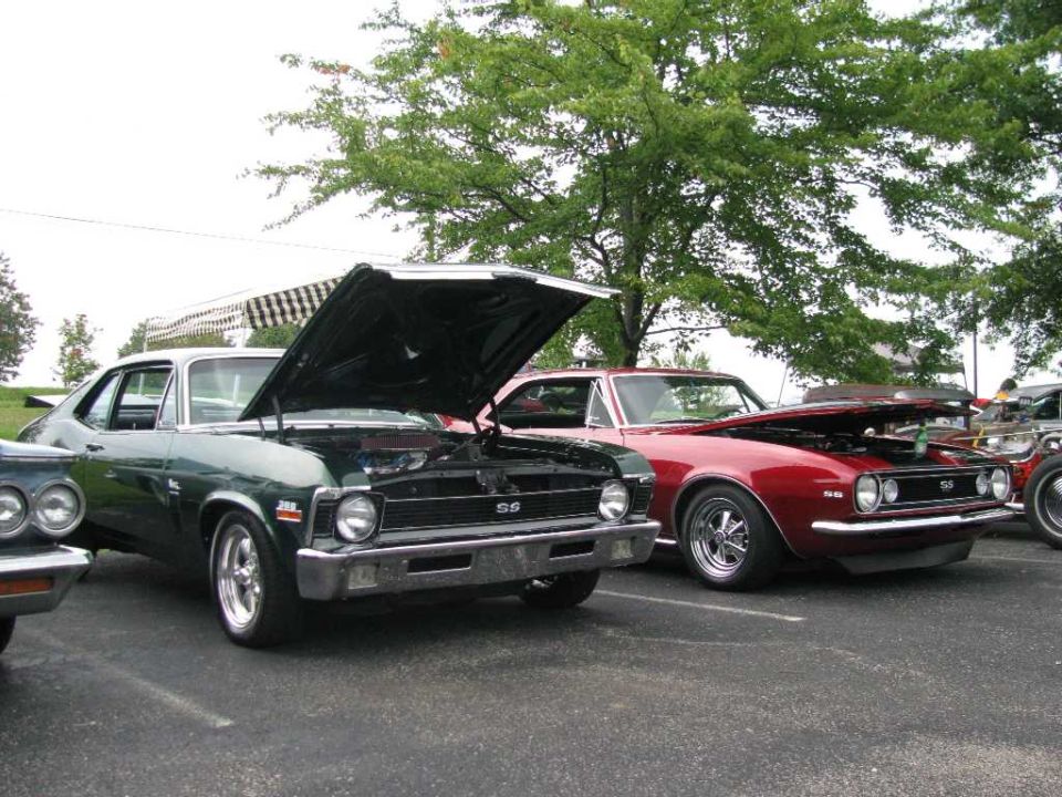 Two classic muscle cars, a dark green one and a red one, parked with hoods open.
