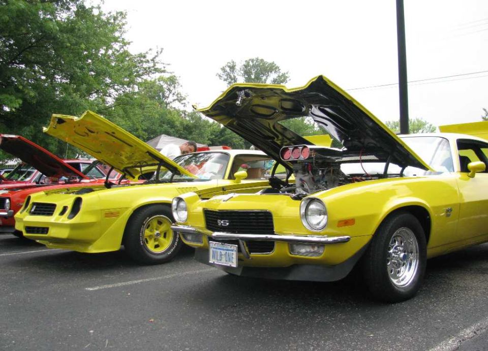 Two yellow classic muscle cars with hoods open, one with a supercharger.