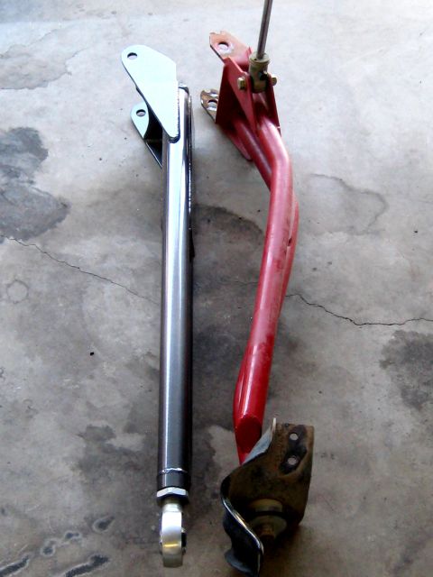1996 Ford Bronco - Two automotive suspension control arms, one silver and one red, are displayed on a concrete surface.