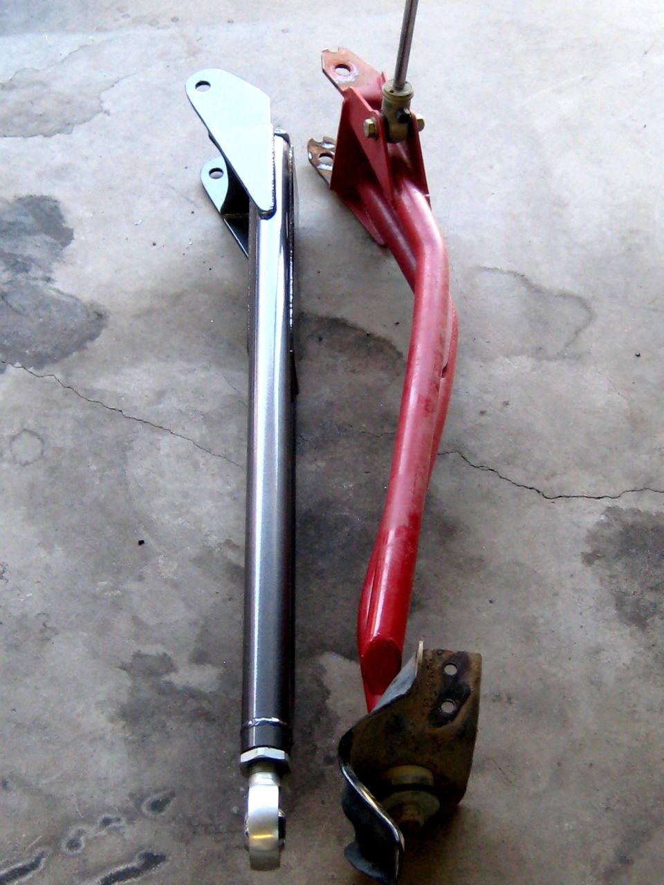 Two suspension control arms, one silver and one red, lie on a concrete floor