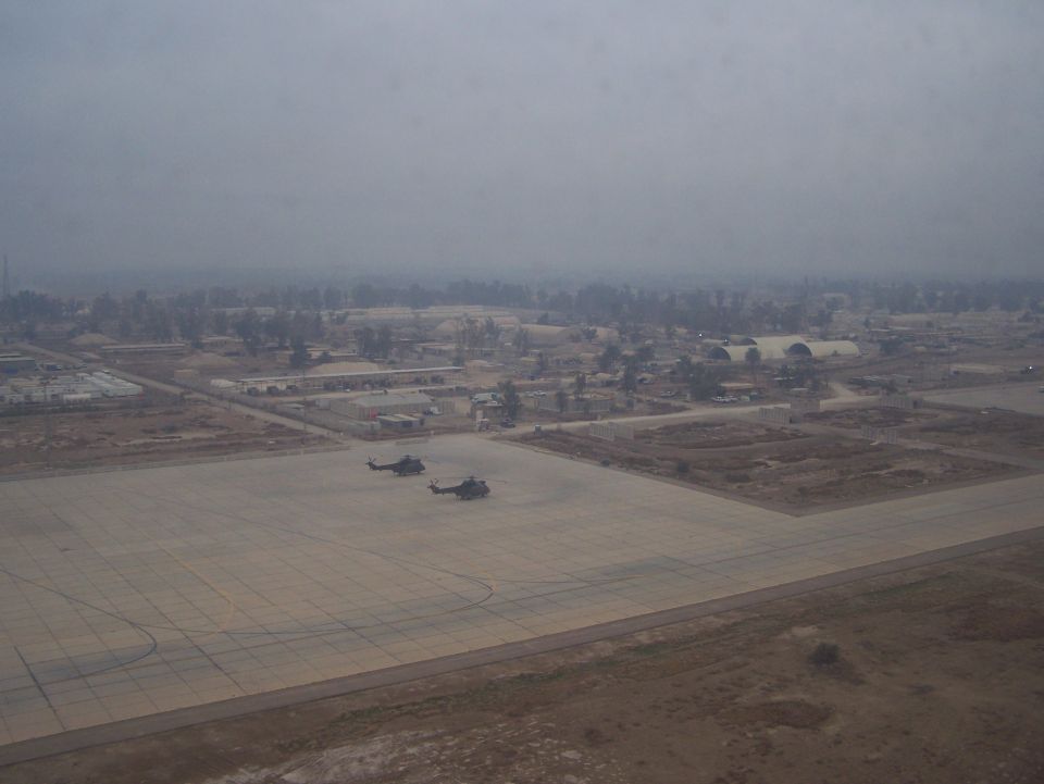 Two helicopters sit on a large concrete airfield with buildings and trees in the background