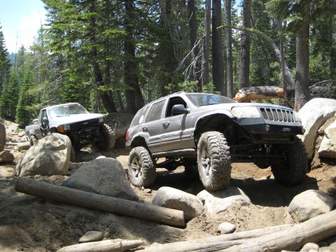 1999 Jeep Grand Cherokee - Two off-road vehicles tackle a challenging, boulder-strewn path surrounded by tall trees.