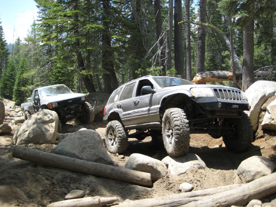 Silver Jeep Grand Cherokee and white Toyota pickup truck navigating a rocky trail in a forest