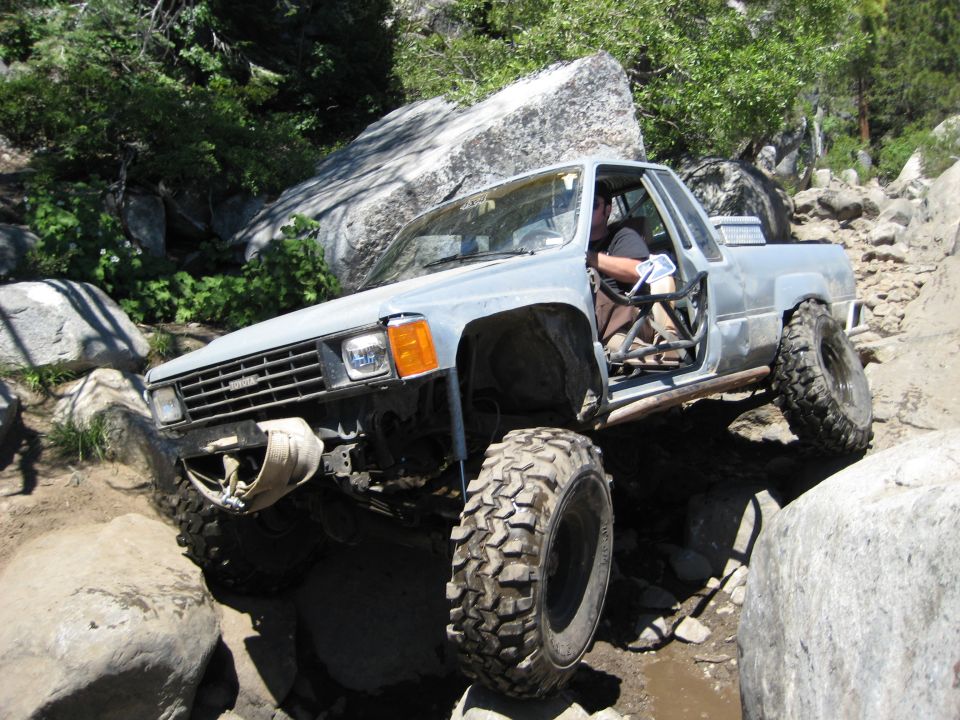 Modified pickup truck with large tires navigating a rocky trail, driver visible inside
