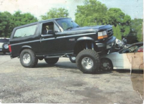 1995 Ford Bronco - A black Ford Bronco is shown with its front end on top of a white car.