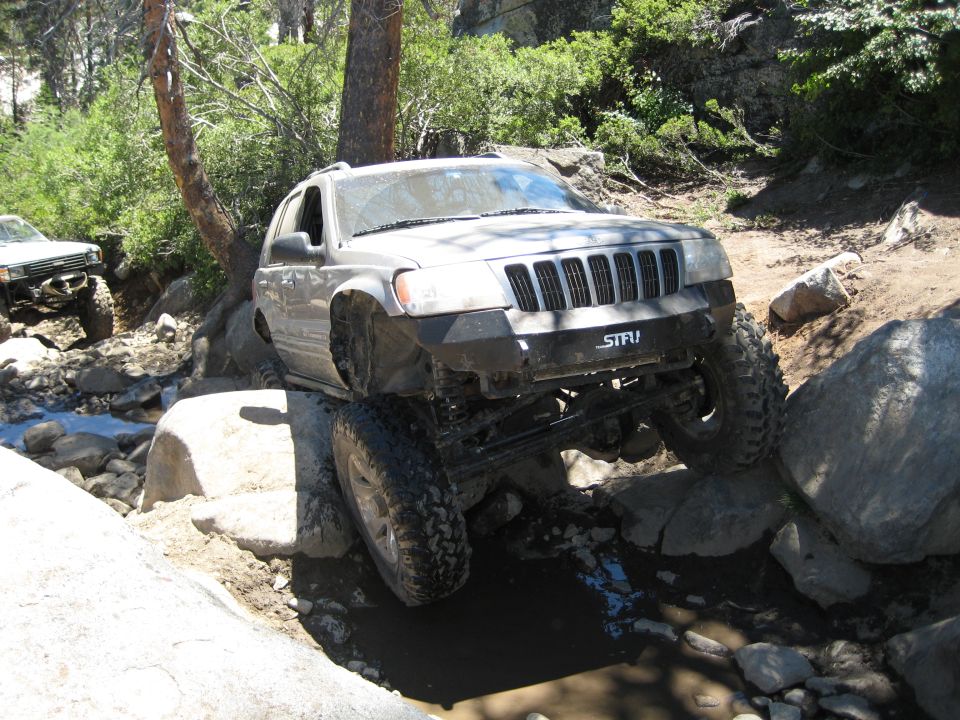 Silver Jeep Grand Cherokee navigating rocks on a trail. Another off-road vehicle is visible behind it
