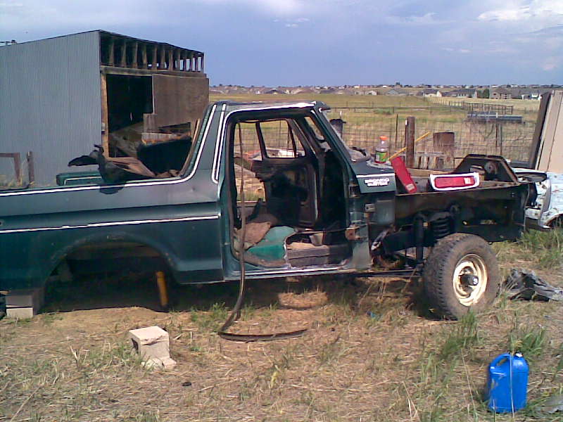 Stripped dark green Ford truck body in a field with front clip and doors removed