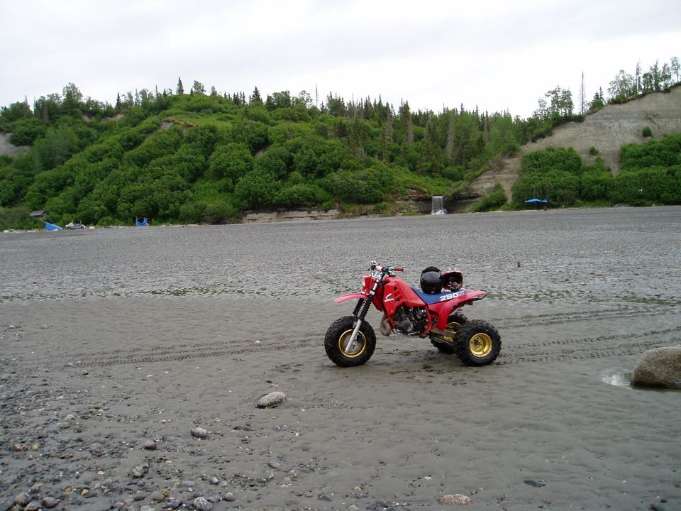Red three-wheeled ATV parked on a gravelly beach with a wooded hill in the background