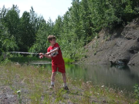 1978 Ford Bronco - A young boy casts his fishing line into a calm lake surrounded by lush green trees and a embankment.