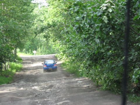 1978 Ford Bronco - A blue sedan navigates a rough dirt road through a wooded area.