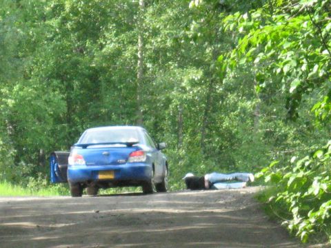 1978 Ford Bronco - A blue sedan is parked on a dirt road with its driver's side door open, and a person is lying on the ground beside it.