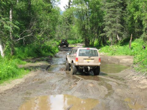 1978 Ford Bronco - Two SUVs navigate a waterlogged dirt road surrounded by dense green forest.