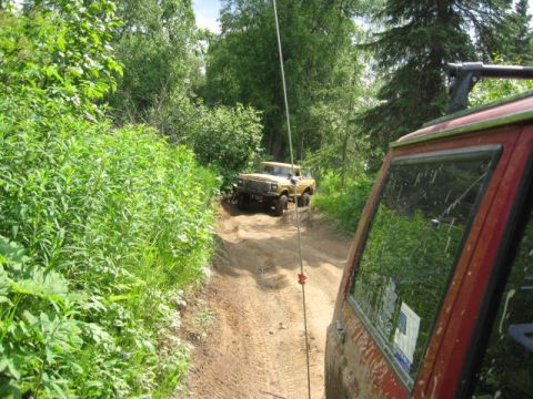 1978 Ford Bronco - Two off-road vehicles navigate a narrow dirt path surrounded by dense green foliage.
