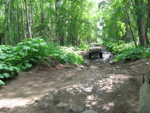 1978 Ford Bronco - A tan off-road vehicle navigates a challenging, muddy, and rocky path through a dense forest.