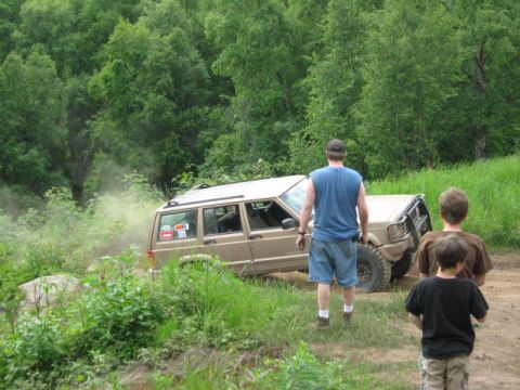 1978 Ford Bronco - A tan SUV navigates a dusty trail surrounded by lush green trees.