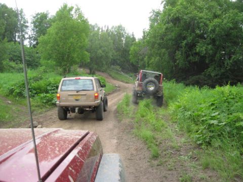 1978 Ford Bronco - Two off-road vehicles navigate a dirt path surrounded by lush green trees and vegetation.