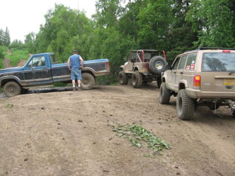 1978 Ford Bronco - Three off-road vehicles navigate a dirt path surrounded by trees.