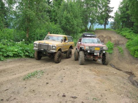 1978 Ford Bronco - Two modified SUVs navigate a steep, muddy off-road trail surrounded by lush green trees.