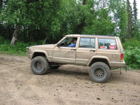 1978 Ford Bronco - A tan Jeep Cherokee XJ is parked on a dirt road surrounded by trees.