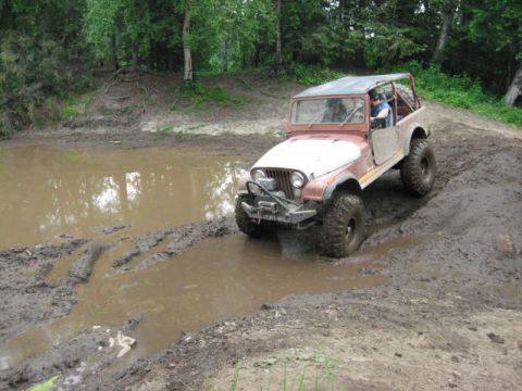 1978 Ford Bronco - A Jeep CJ-7 is driven through a muddy obstacle course with trees in the background.