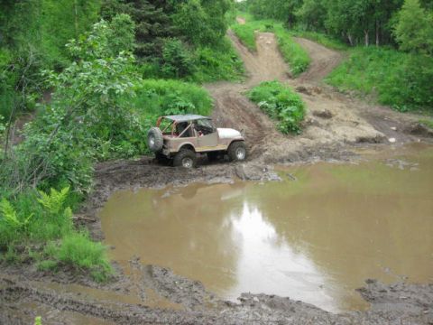 1978 Ford Bronco - An off-road vehicle navigates a muddy trail, with a large puddle reflecting the sky and surrounding trees.