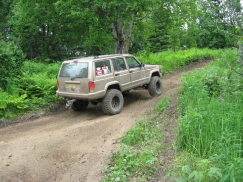 1978 Ford Bronco - A tan Jeep Cherokee XJ navigates a dirt path surrounded by lush green foliage and trees.