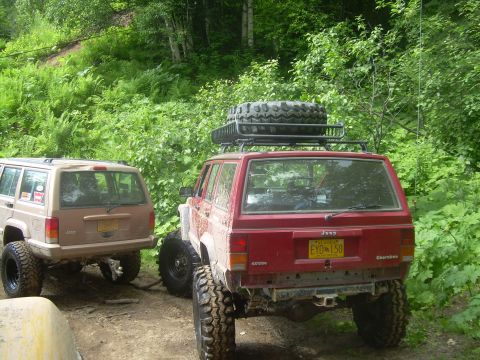 1978 Ford Bronco - Two modified Jeep Cherokees are parked on a dirt path surrounded by lush green foliage.