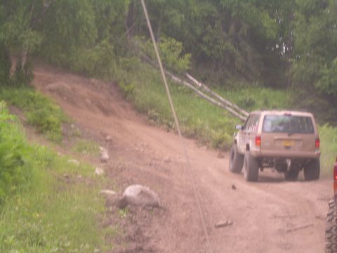1978 Ford Bronco - A tan SUV navigates a dirt path surrounded by greenery, with another vehicle partially visible in the foreground.