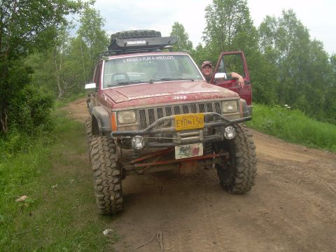 1978 Ford Bronco - Modified red Jeep Cherokee XJ parked on a dirt road with trees in the background.