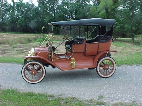 1912 Ford Model T - Driver-side profile of a brass-era open touring car painted rust red, parked on a gravel drive with grass and trees behind. It has a black…