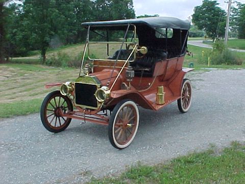 1912 Ford Model T - Rust-red brass-era open touring car shot from the front three-quarter on a gravel drive with a grassy hillside behind. Brass trim is promin…