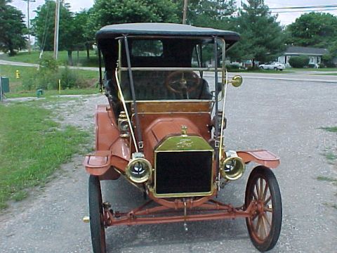 1912 Ford Model T - Head-on front view of a rust-red brass-era open touring car on a gravel shoulder, showing the brass-framed radiator with black core, two br…