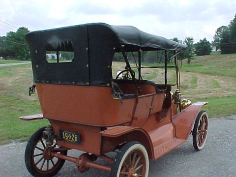 1912 Ford Model T - Rust-red brass-era open touring car photographed from the rear three-quarter on a paved drive beside a mown field. It wears a black folding…