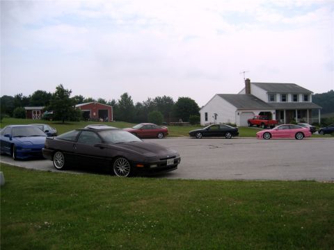 1993 Ford Probe - Several coupes parked on a paved lot by a white house and red barn, foreground dark Ford Probe, pink and blue cars beyond.