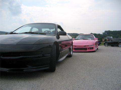 1993 Ford Probe - A black Ford Probe is parked in the foreground, angled towards the viewer. A pink Ford Probe is parked behind it, with other vehicles visib…