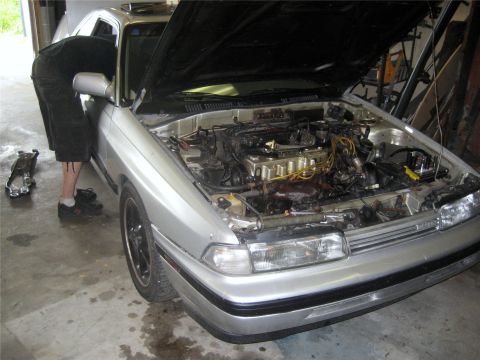 1993 Ford Probe - A person in black shorts and a black t-shirt works on the engine of a silver coupe with the hood open. The engine bay is exposed, showing v…