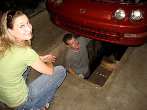 1993 Ford Probe - A woman in a green shirt and jeans smiles at the camera while a man works on a red car that is raised over a pit.