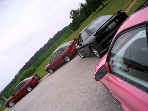 1993 Ford Probe - Four cars are parked diagonally on an asphalt road with a grassy hillside and trees in the background. The closest car is pink, followed by…