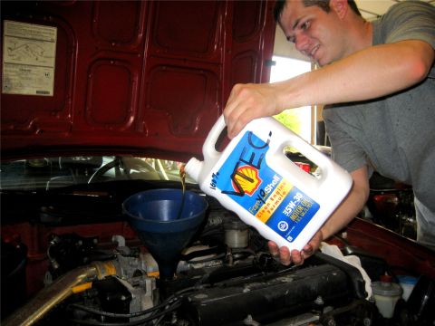 1993 Ford Probe - A man pours motor oil from a white jug into a blue funnel placed in the engine bay of a red car.
