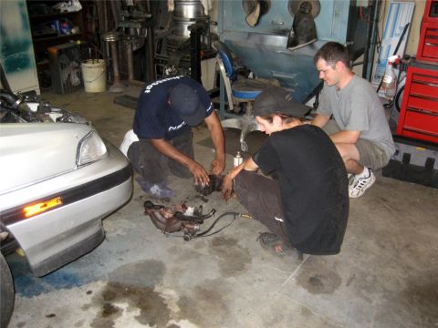 1993 Ford Probe - Three men work on a car engine disassembled on the floor of a garage. The front of a silver car is visible on the left.