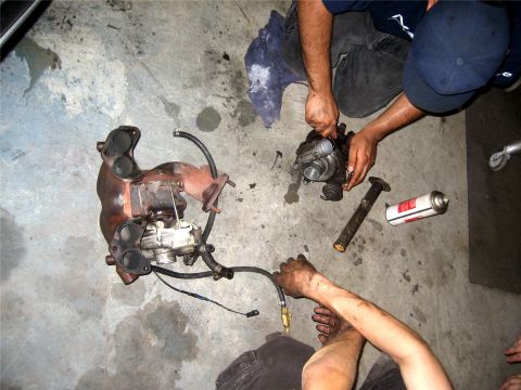 1993 Ford Probe - A turbocharger and exhaust manifold are laid out on a concrete floor next to a can of spray lubricant and a metal pipe. Two people are work…