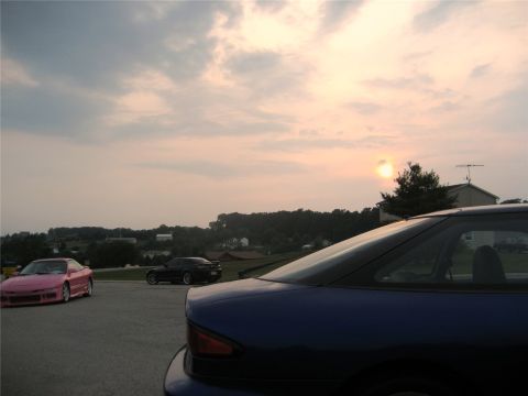 1993 Ford Probe - A pink Ford Probe is parked in a lot with a dark blue car in the foreground and a dark car in the background. The sun is setting behind a t…