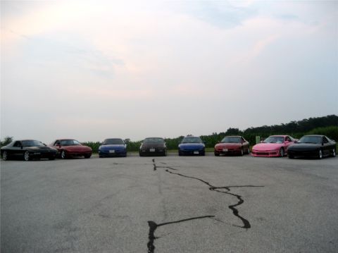 1993 Ford Probe - A line of seven sports coupes are parked on asphalt in front of a field of crops under a cloudy sky. The cars are of various colors includi…