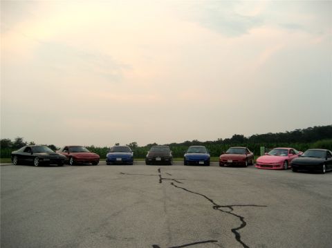 1993 Ford Probe - A line of seven sports coupes are parked on asphalt in front of a tree line under a hazy sky. The cars are various colors including black,…