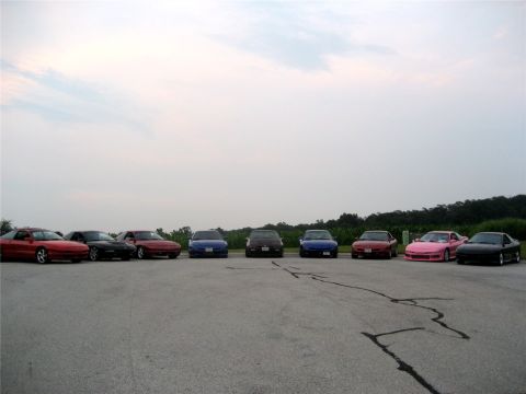 1993 Ford Probe - A line of eight sports cars are parked in a lot with a tree line in the background under a cloudy sky. The cars are various colors includin…
